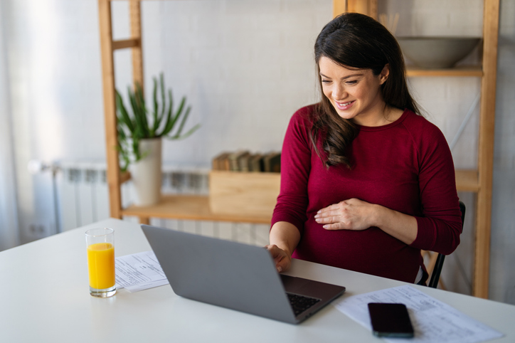 vrouw zit aan laptop met hand op zwangere buik en glas fruitsap op tafel